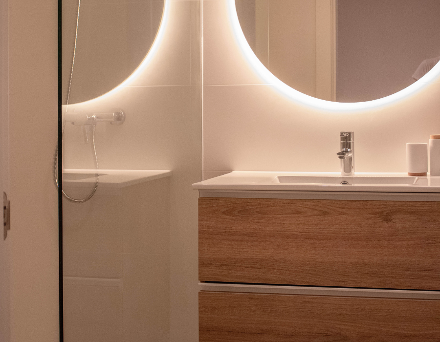 Έπιπλο μπάνιου σε χρώμα ξύλου με 2 συρτάρια και ενσωματωμένο λευκό νιπτήρα πορσελάνης. Inox βρύση νιπτήρα και led καθρέφτης μπάνιου. Αριστερά φαίνεται μέρος του ντουζ με κυρίαρχο το προστατευτικό τζάμι ντουζ. Το μπάνιο έχει λευκό πλακάκι στους τοίχους.