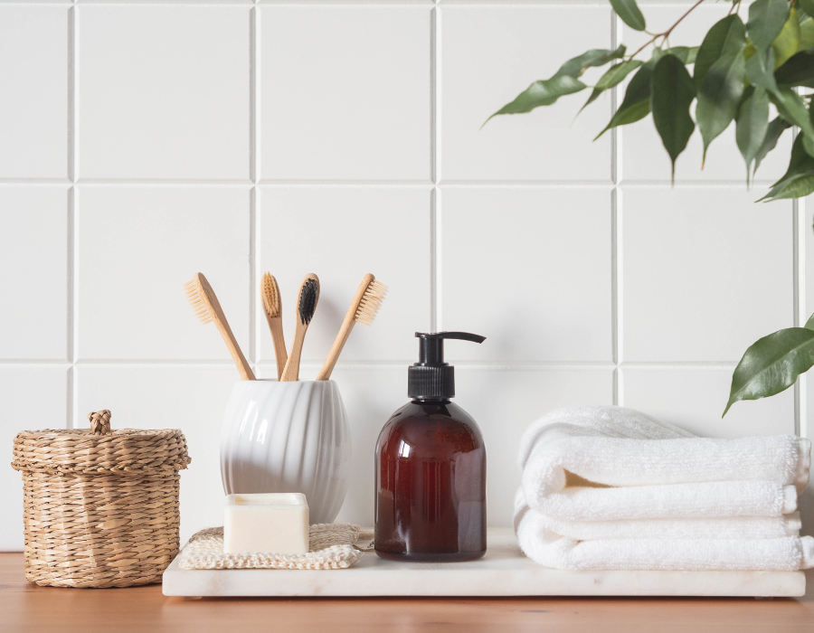 Είδη ενός οικολογικού μπάνιου μπροστά από τετράγωνο λευκό πλακάκι. Οδοντόβουρτσες και μικρό καλάθι αποθήκευσης από bamboo, γυάλινο δοχείο κρεμοσάπουνου, λευκές πετσέτες μπάνιου από βαμβάκι.